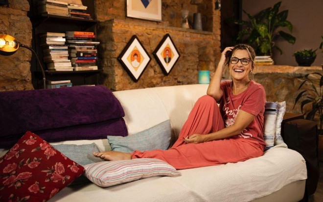Ingrid Guimarães sentada em um sofá com uma das pernas esticadas. Há objetos na parede