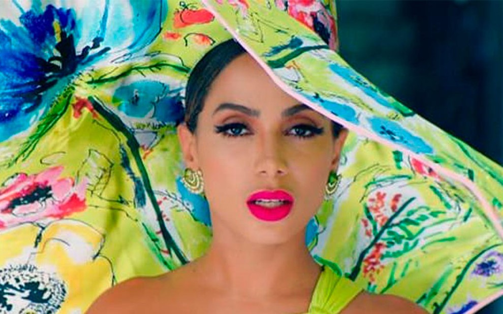Anitta em cena do clipe Me Gusta: cantora usa batom rosa choque, está com o maiô verde limão e chapéu estampado