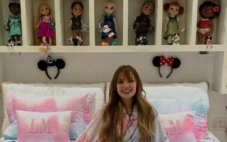 A atriz Larissa Manoela posa em cima de sua cama com uma coleção de bonecas em estante pendurada em seu quarto; foto publicada nas redes sociais