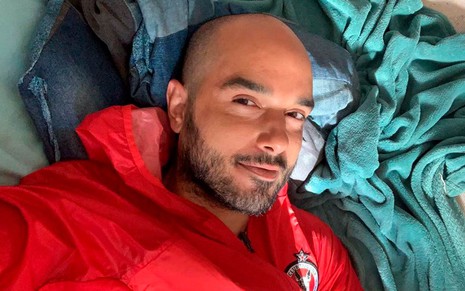 Leo Rosa em foto publicada no Instagram: deitado, ator sorri para a câmera e usa moletom vermelho