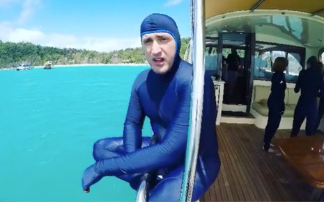 Paulo Gustavo com roupa de mergulho azul, ele está sentado na lateral do barco