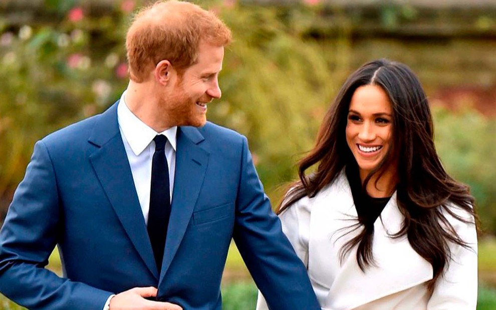 O príncipe Harry e a atriz Meghan Markle em anúncio do noivado: ele usa terno azul, gravata preta e olha para a atriz, que está de casaco branco e olha com felicidade para fora do quadro