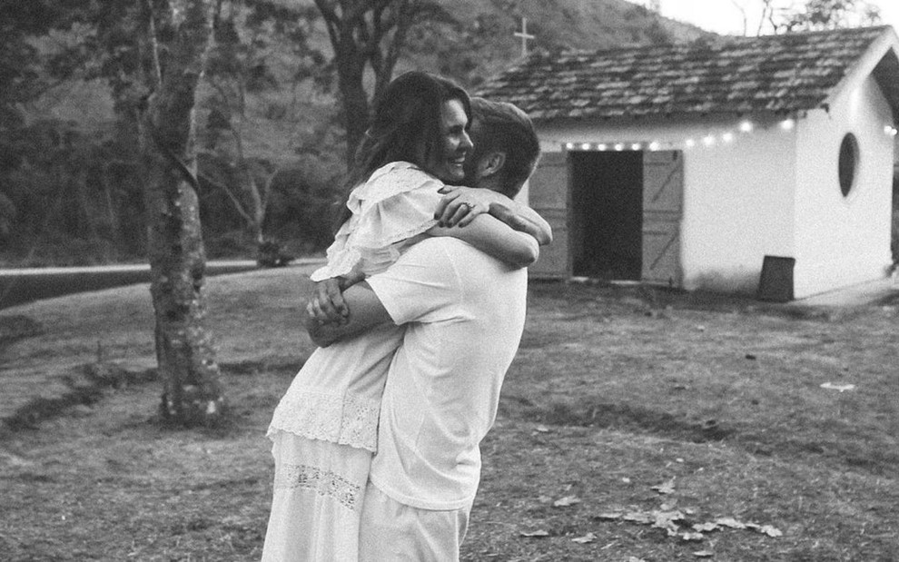 O casal Fernanda Lima e Rodrigo Hilbert em foto preto e branco em que aparecem abraçados em frente à capela do sítio da família