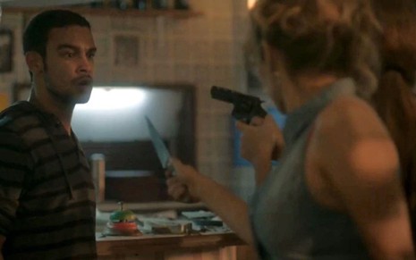 Acuado e com expressão de medo, Jacaré (Sergio Malheiros) observa Eliza (Marina Ruy Barbosa) com uma arma e Gilda (Leona Cavalli) com um facão em cena de Totalmente Demais
