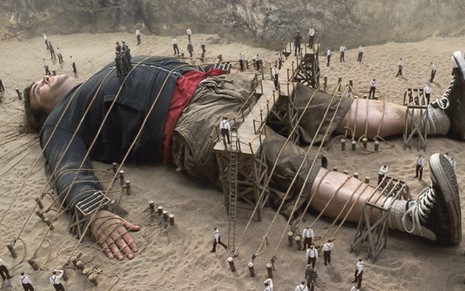 O ator Jack Black (Lemuel Gulliver) jogado no chão e amarrado por várias cordas; ele está rodeado de pessoas muito pequenas
