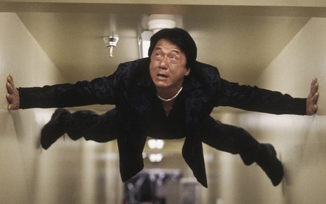 Jackie Chan com expressão assustada em A Hora do Rush 2 (2001); ele se segura entre duas paredes em pose de espião