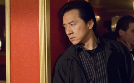 Jackie Chan com expressão séria em A Hora Do Rush 3 (2007); ele tenta espiar um ambiente por trás da parede