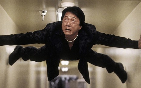 o ator Jackie Chan se segura em uma parede com auxílio das pernas e dos braços com expressão de sofrimento
