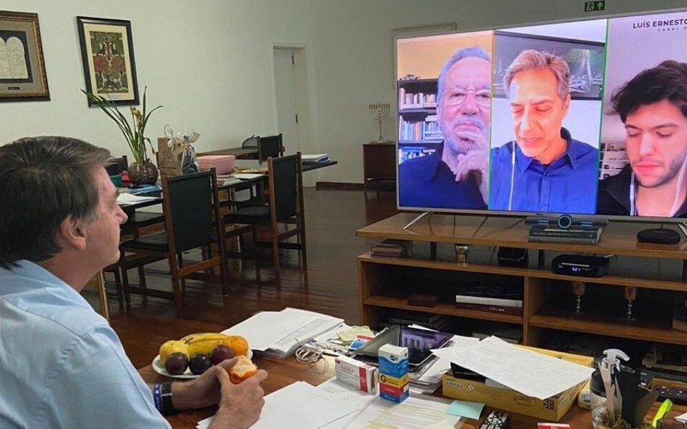 Imagem de Jair Bolsonaro, de costas, vendo televisão com vídeo de Alexandre Garcia, Luís Ernesto Lacombe e Caio Coppolla na TV