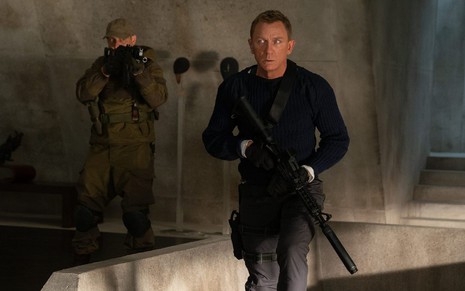 James Bond (Daniel Craig) de roupa especial preta segurando uma arma no lado direito e homem no lado esquerdo mirando uma arma para sua cabeça