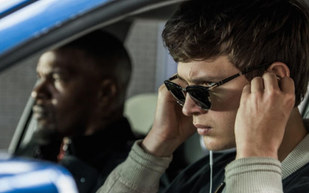 Jamie Foxx (ao fundo) e Ansel Elgort (de óculos escuros e colocando fone de ouvido) estão dentro de um carro em cena de Em Ritmo de Fuga