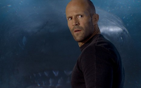 Jason Statham está com expressão de surpresa; ele aparece em frente a um vidro que reflete um tubarão gigante em cena de Megatubarão (2018)