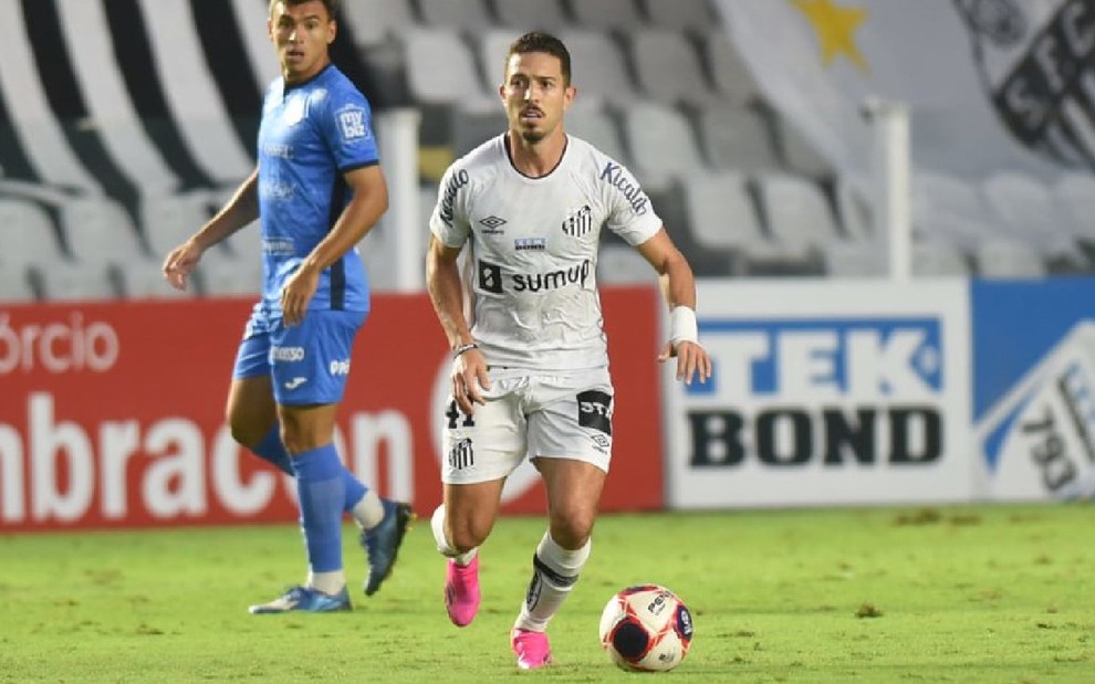 Jean Mota com o uniforme todo branco do Santos conduz bola sendo observado por adversário de uniforme azul