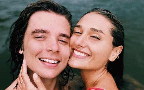 João Figueiredo e Sasha Meneghel sorridentes, de rostos colados, dentro do mar