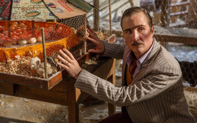 O ator João Miguel caracterizado de Ernesto, personagem do filme Quase Memória, vestido com terno bege listrado com cinza e camisa azul claro, agachado com as mãos em uma gaiola com pintinhos e ovos