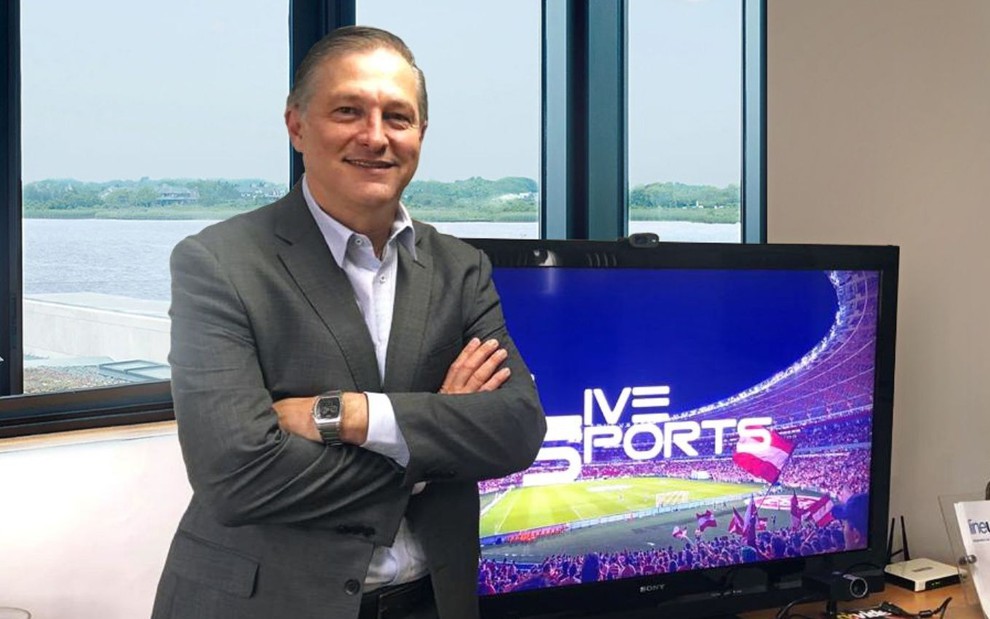 O jornalista João Palomino de braços cruzados ao lado de televisão com o logo da LiveSports