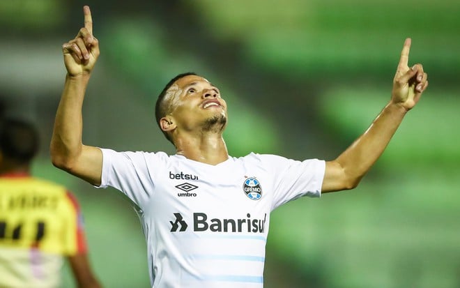 Darlan com camisa branca do Grêmio com os braços abertos e com os dois indicadores apontados para cima