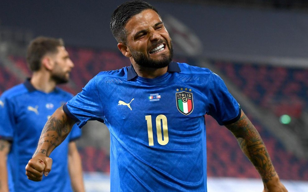 Lorenzo Insigne com camisa azul da Itália com punho da mão direita cerrado e sorriso no rosto com outro jogador italiano ao fundo desfocado