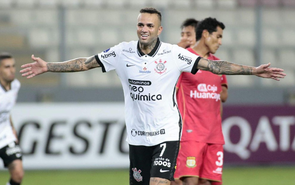 Luan com a camisa branca do Corinthians celebra com braços abertos e sorriso no rosto com adversários de camisa vermelha ao fundo