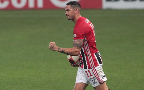 Luciano com camisa vermelha, branca e preta do São Paulo e calção branco segurando a bola palma da mão direita e cerrando a mão esquerda em vibração