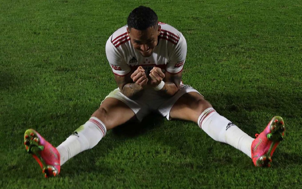 Luciano com uniforme do São Paulo sentado no gramado com as pernas abertas e os dois punhos juntos em comemoração