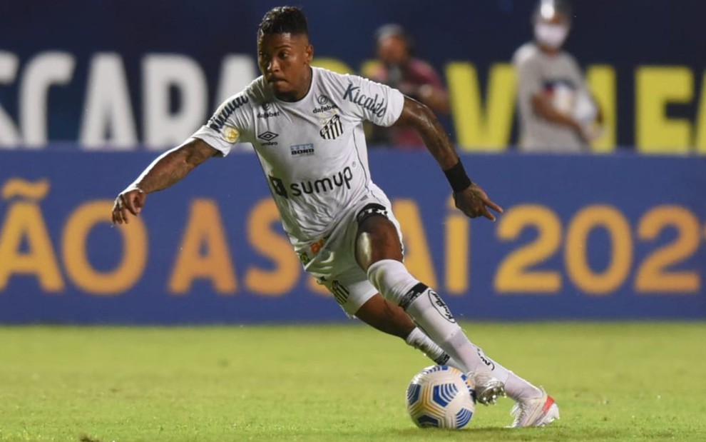 Marinho com o uniforme todo branco do Santos correndo em campo com a bola no pé esquerdo e braços abertos