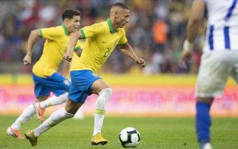 Richarlison com a camisa amarela, calção azul e meia branca da Seleção Brasileira corre com a bola em direção à direita com outro jogador do Brasil ao fundo