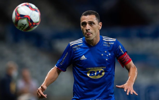 Rômulo com uniforme azul do Cruzeiro com a língua para fora e braços abertos olhando para a bola