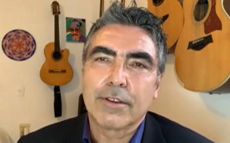 John olha para frente, usa terno preto e camiseta azul, ele está na frente de uma parede branca com violões pendurados