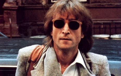 John Lennon de óculos escuros, camisa branca e paletó cinza olhando para a câmera