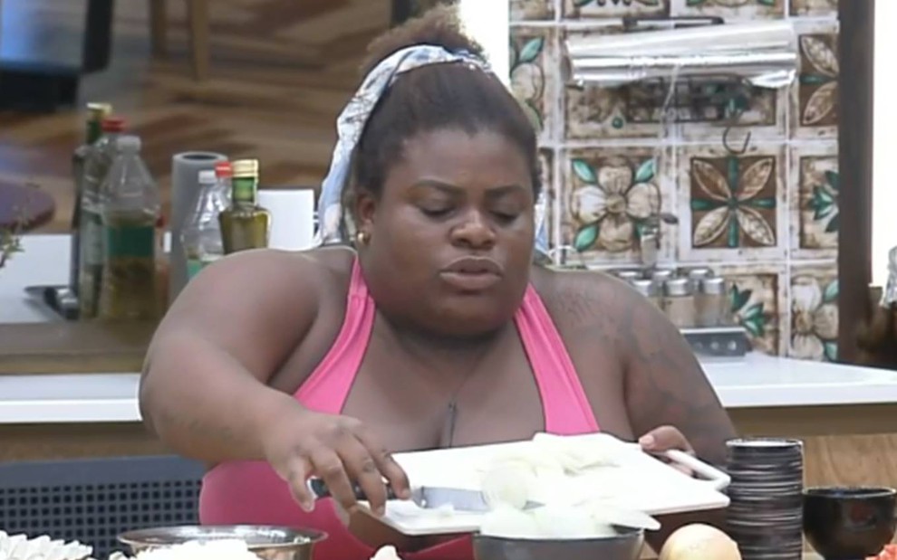 Jojo aparece com um lenço no cabelo, usa regata cor de roça e está sentada preparando o almoço em A Fazenda 12