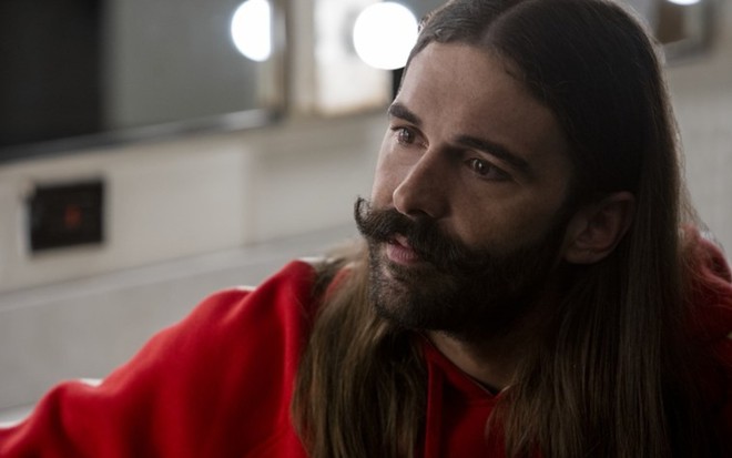 Jonathan Van Ness no lado direito com blusa vermelha e cabelos soltos sobre o ombro e grande bigode penteado