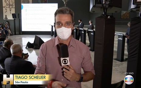 O repórter Tiago Scheuer aparece de máscara e segurando um microfone durante a coletiva de imprensa de João Doria (ao fundo) na quarta-feira (3) na Globo