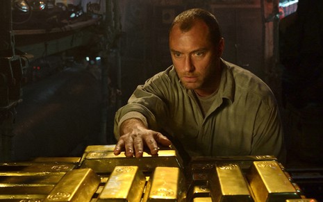 Jude Law num lugar escuro com a mão em cima de barras de ouro