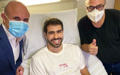 Juliano Laham de camiseta branca, deitado em cama de hospital, ao lado de dois médicos