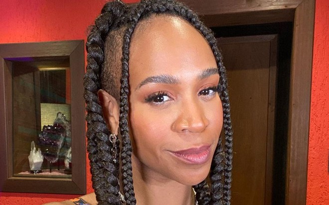 Karol usa tranças no cabelo, olha para a câmera e está com uma blusa de alça azul e verde