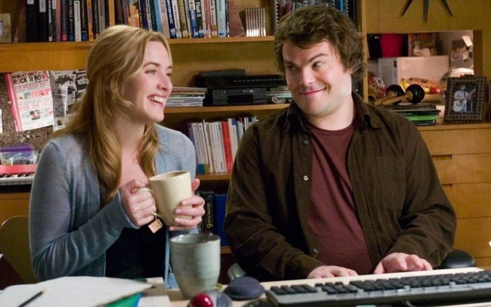 Kate Winslet e Jack Black sorriem em cena do filme O Amor Não Tira Férias (2006); ela segura uma caneca, e ele toca um teclado