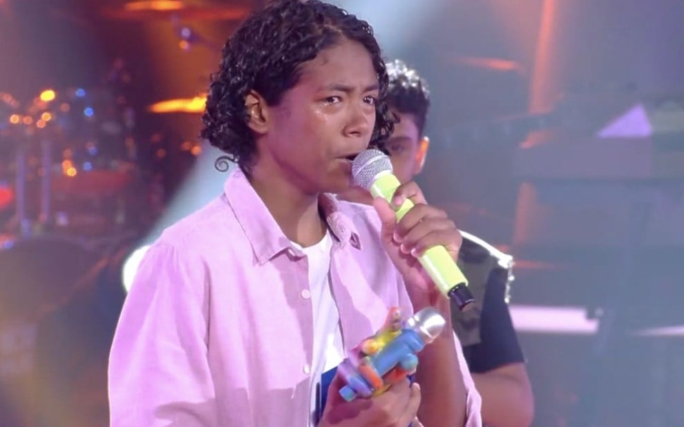 Kauê Penna chorando no palco do The Voice Kids, usando camisa rosa