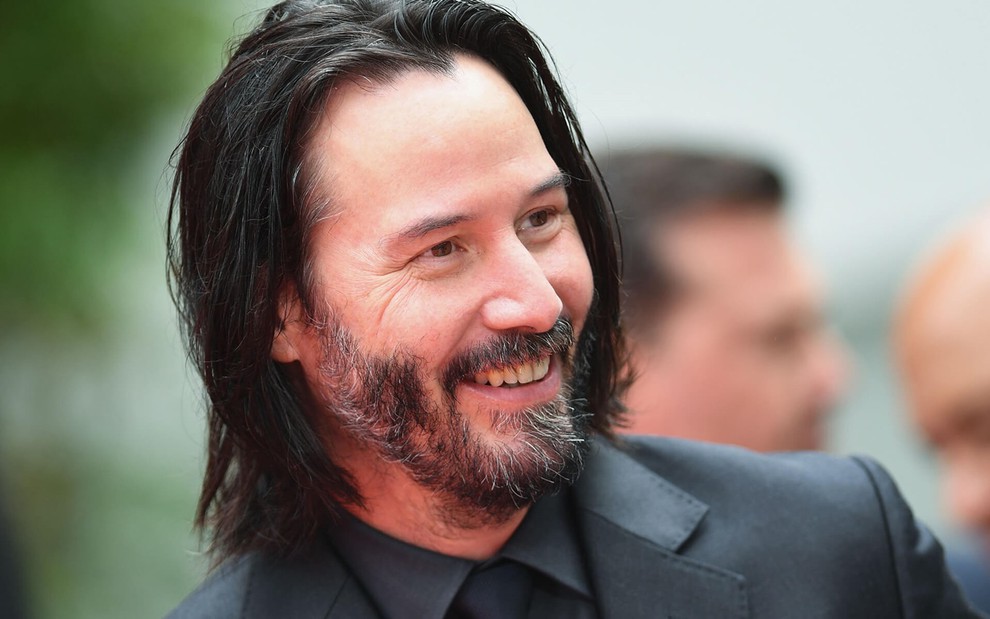 Keanu Reeves usa um terno preto e sorri para a foto em tapete vermelho