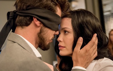 O ator Bruno Ferrari de olhos vendados encosta a testa na da atriz Vitória Strada, que usa uma peruca, em cena como Rafael e Kyra de Salve-se Quem Puder