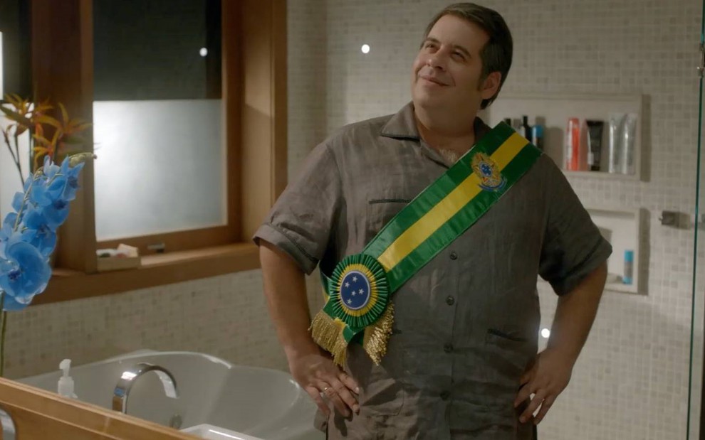 Leandro Hassum se olhando no espelho do banheiro, com as mãos na cintura, usando uma faixa do Brasil no corpo