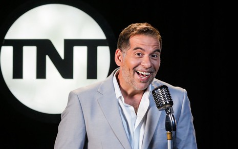 Leandro Hassum sorrindo enquanto posa para foto na frente de um microfone; ao fundo, o logo da TNT