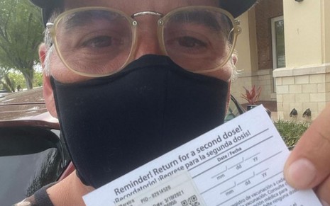 Leandro Hassum, de máscara, óculos e boné, mostra comprovante de que tomou a vacina nos EUA
