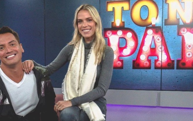 Leo Dias e Ligia Mendes sorrindo, lado a lado, em estúdio da rádio Jovem Pan, com Tô na Pan escrito num telão ao fundo