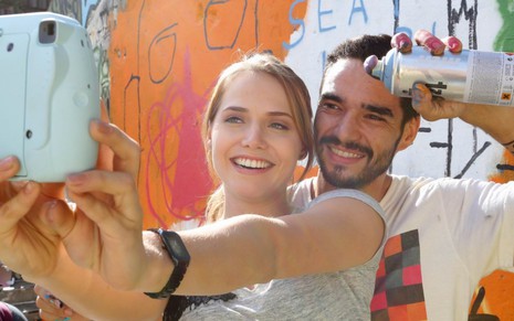 Leticia Colin segura uma câmera azul e tira selfie sorrindo com Caio Blat, que está com uma tinta spray na mão e também sorri