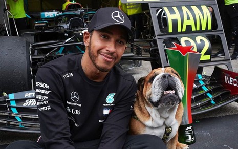 Lewis Hamilton com uniforme da Mercedez, boné preto, ao lado de seu cão de estimação