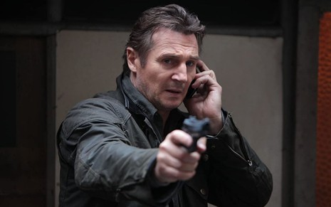 Liam Neeson fala no celular enquanto aponta uma arma para frente em Busca Implacável 2 (2012)