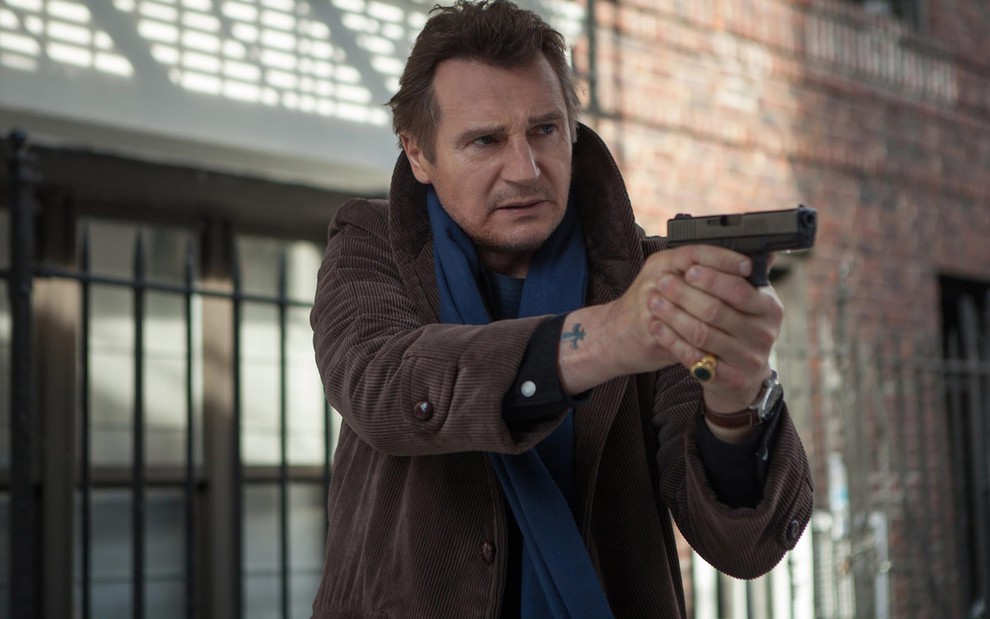 Liam Neeson usa jaqueta marrom de riscas, cachecol azul e empunha uma arma