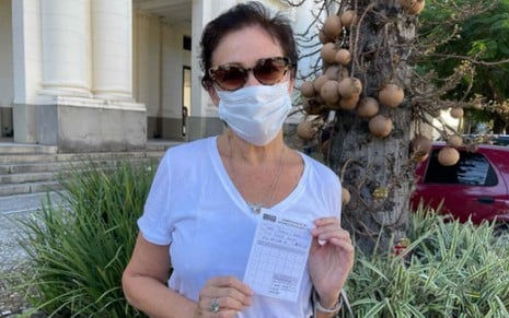 Lilia Cabral de camiseta e máscara brancas, óculos escuros, segurando a caderneta de vacinação
