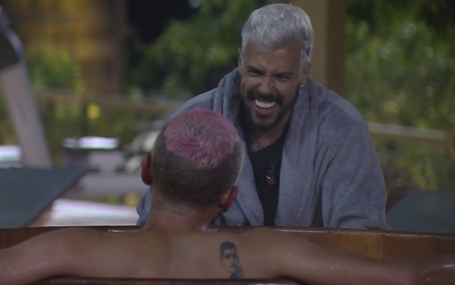 Lipe Ribeiro vestindo um roupão e rindo enquanto conversa com Lucas Selfie, de costas